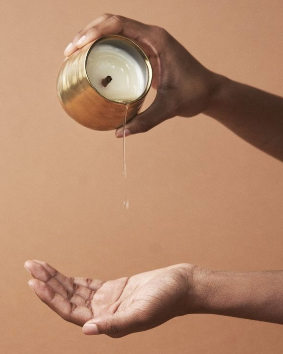 Hand pouring melted soy wax from gold container above open palm over beige background